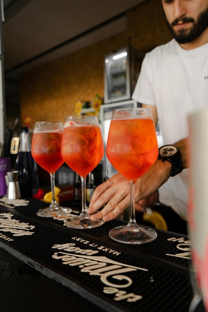 Three Aperol Spritz cocktails on bar counter with bartender preparing drinks.