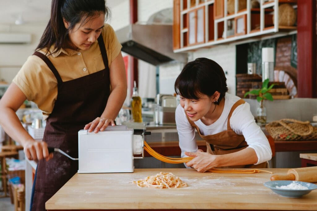 Two Asian women using a pasta machine in a home kitchen setting.
