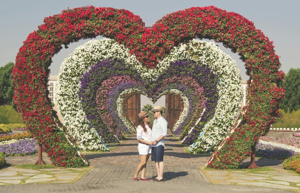 A couple embracing in a vibrant flower garden in Dubai, featuring heart-shaped floral arches.