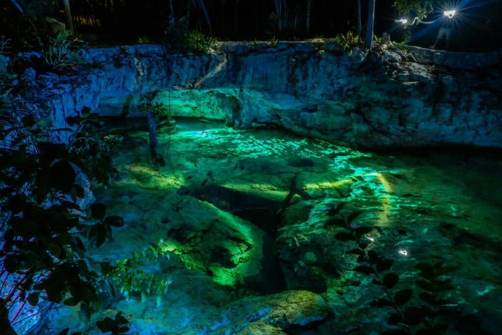Vibrant night view of Grand Cenote in Tulum, Mexico, highlighting illuminated turquoise waters and rocky surroundings.
